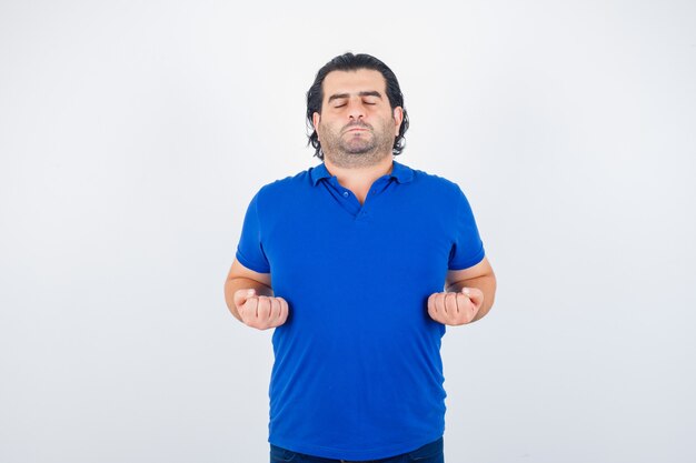 Hombre maduro meditando, manteniendo los ojos cerrados en camiseta azul, jeans y mirando tranquilo, vista frontal.