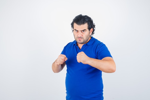 Hombre maduro de pie en pose de lucha en camiseta azul, jeans y mirando confiado. vista frontal.