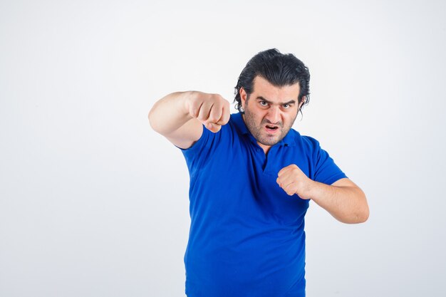 Hombre maduro de pie en pose de lucha en camiseta azul, jeans y mirando confiado. vista frontal.