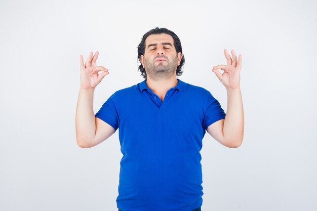 Hombre maduro de pie en pose de meditación en camiseta azul, jeans y mirando relajado, vista frontal.