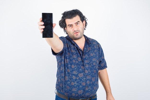Foto gratuita hombre maduro que estira la mano para mostrar el teléfono móvil en camisa y parece seguro. vista frontal.