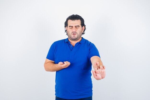 Hombre maduro que muestra el gesto de la pistola, estirando la mano mientras sostiene algo en una camiseta azul, jeans y parece tranquilo. vista frontal.