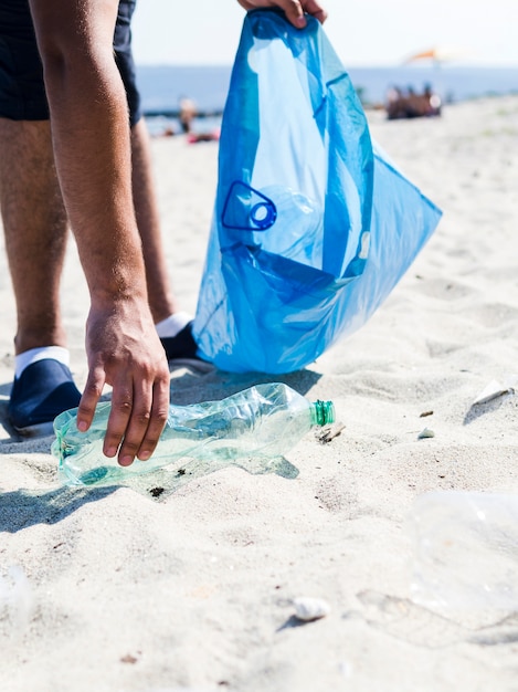 Hombre mano recogiendo basura botella de plástico en la playa mientras sostiene la bolsa de basura azul