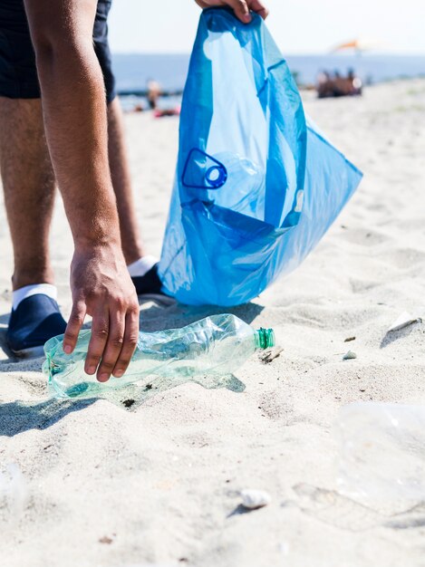 Hombre mano recogiendo basura botella de plástico en la playa mientras sostiene la bolsa de basura azul