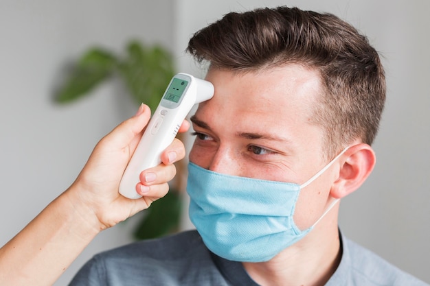 Hombre con máscara médica comprobando la temperatura en la oficina con termómetro