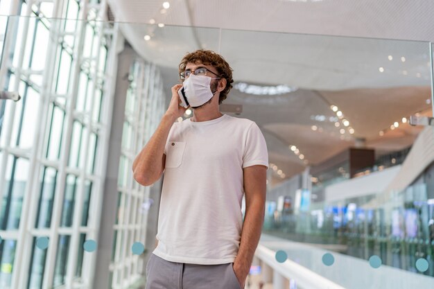 Hombre con máscara protectora de pie en el pasillo del aeropuerto con smartphone