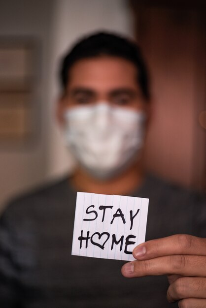 Un hombre con una mascarilla y sosteniendo un trozo de papel con un escrito '' Quédate en casa ''