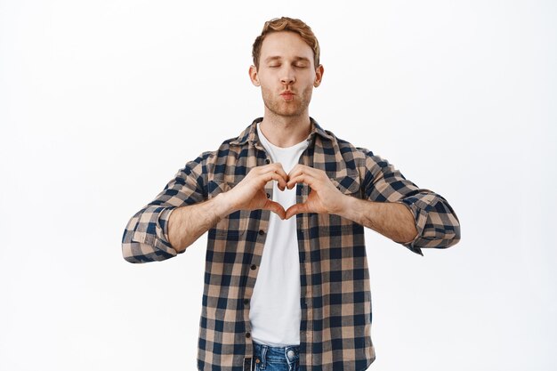 Hombre masculino pelirrojo lindo y tonto que muestra el signo del corazón, dice te amo, cierra los ojos y espera un beso con los labios fruncidos, de pie contra la pared blanca