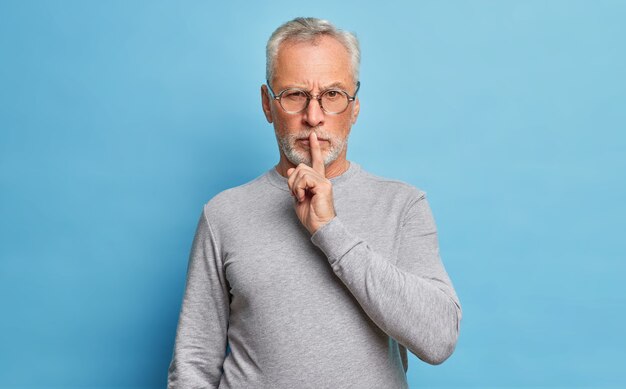 Hombre mayor barbudo tiene una expresión seria, hace un gesto de silencio, pide silencio con el dedo en los labios, exige silencio, usa anteojos ópticos y un jersey de manga larga aislado en una pared azul