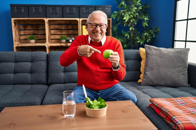 Foto gratuita hombre mayor con cabello gris comiendo ensalada y manzana verde sonriendo feliz señalando con la mano y el dedo