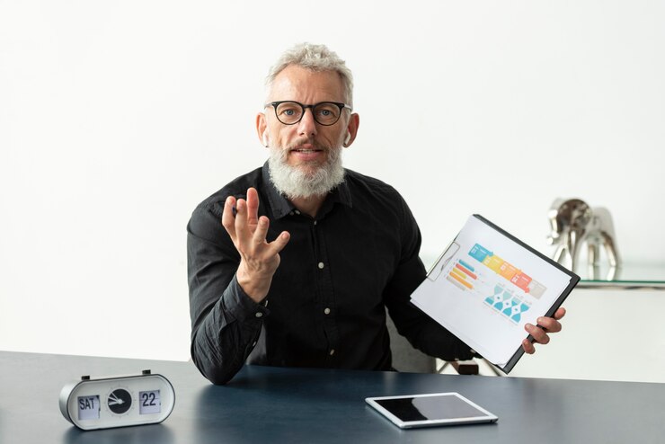 Foto gratuito hombre mayor en casa que muestra el gráfico en el bloc de notas con tableta en el escritorio