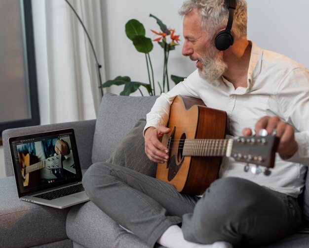 Hombre mayor en casa en el sofá usando la computadora portátil para estudiar lecciones de guitarra