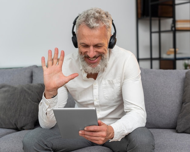 Hombre mayor en casa en el sofá con una videollamada en tableta y usando audífonos