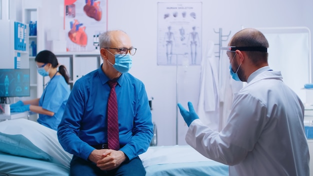 Hombre mayor en cita médica durante la pandemia de COVID-19. Paciente con máscara y médico con ropa protectora. Consulta sanitaria, sistema medicinal. Clínica privada moderna