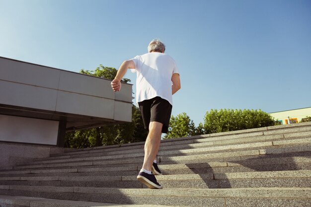 Hombre mayor como corredor con brazalete o rastreador de fitness en la calle de la ciudad. Modelo masculino caucásico practicando jogging y entrenamientos de cardio en la mañana de verano. Estilo de vida saludable, deporte, concepto de actividad.