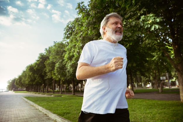 Hombre mayor como corredor con brazalete o rastreador de fitness en la calle de la ciudad. Modelo masculino caucásico que practica jogging y entrenamientos de cardio en la mañana de verano. Estilo de vida saludable, deporte, concepto de actividad.