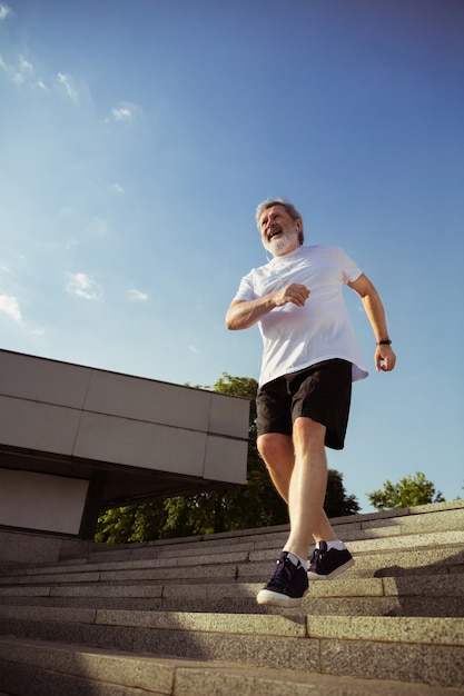 Hombre mayor como corredor con brazalete o rastreador de fitness en la calle de la ciudad. Modelo masculino caucásico que practica jogging y entrenamientos de cardio en la mañana de verano. Estilo de vida saludable, deporte, concepto de actividad.