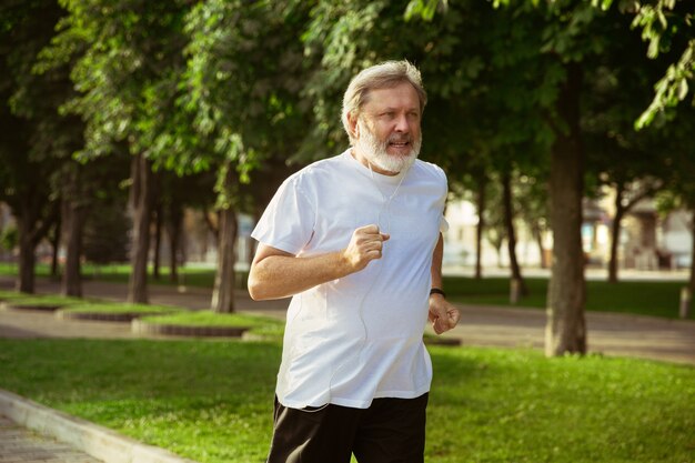 Hombre mayor como corredor con brazalete o rastreador de fitness en la calle de la ciudad. Modelo masculino caucásico que practica jogging y entrenamientos de cardio en la mañana de verano. Estilo de vida saludable, deporte, concepto de actividad.