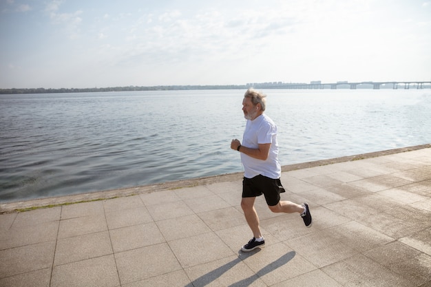 Hombre mayor como corredor con brazalete o rastreador de fitness en la orilla del río. Modelo masculino caucásico que practica jogging y entrenamientos de cardio en la mañana de verano. Estilo de vida saludable, deporte, concepto de actividad.