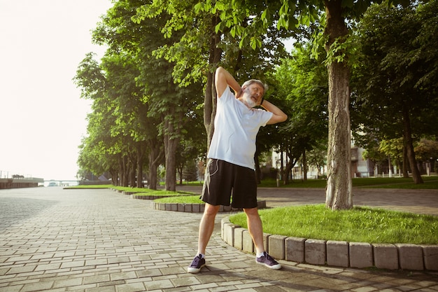 Hombre mayor como corredor en la calle de la ciudad. Modelo masculino caucásico de jogging y entrenamiento cardiovascular en la mañana de verano. Haciendo ejercicios de estiramiento cerca de la pradera. Estilo de vida saludable, deporte, concepto de actividad.