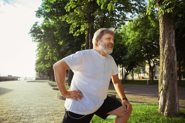 Hombre mayor como corredor en la calle de la ciudad. Modelo masculino caucásico de jogging y entrenamiento cardiovascular en la mañana de verano. Haciendo ejercicios de estiramiento cerca de la pradera. Estilo de vida saludable, deporte, concepto de actividad.