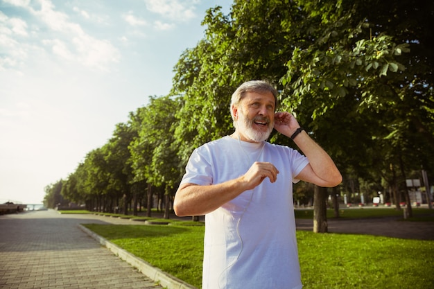 Hombre mayor como corredor con rastreador de fitness en la calle de la ciudad. Modelo masculino caucásico usando gadgets mientras hace jogging y entrenamiento cardiovascular en la mañana de verano. Estilo de vida saludable, deporte, concepto de actividad.