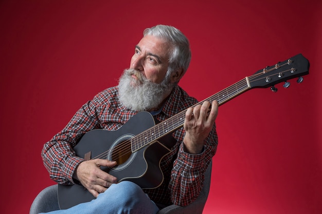 Hombre mayor contemplado que toca la guitarra contra fondo rojo