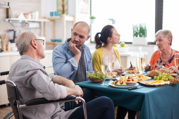 Hombre mayor discapacitado en silla de ruedas que tiene una conversación con su hijo durante el brunch familiar en la cocina. Padres mayores junto con niños maduros.