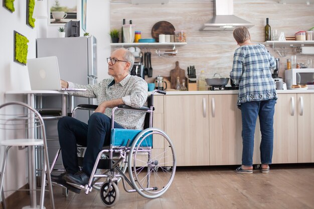 Hombre mayor discapacitado en silla de ruedas trabajando en la computadora portátil en la cocina mientras la esposa está preparando un delicioso desayuno para ambos. Hombre que usa tecnología moderna mientras trabaja desde casa.