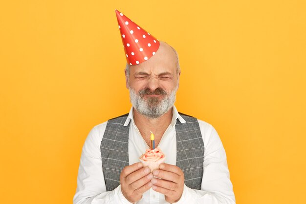 Hombre mayor elegante guapo con barba gris espesa con sombrero de cono cerrando los ojos, pidiendo deseo