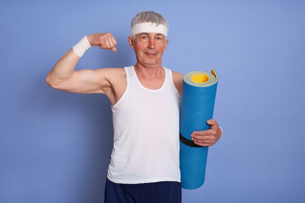 Hombre mayor enérgico tiene entrenamiento físico, sosteniendo una estera de yoga, mostrando bíceps y su poder