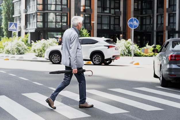 Foto gratuita hombre mayor con estilo en la ciudad cruzando la calle mientras sostiene el paraguas