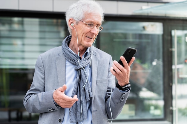Hombre mayor con estilo en la ciudad con teléfono inteligente y auriculares para videollamada