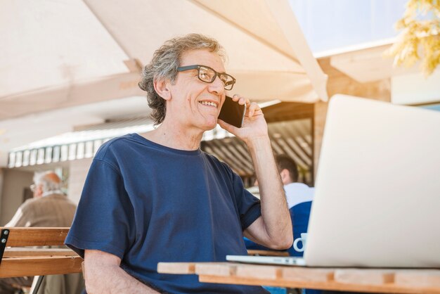 Hombre mayor feliz que habla en el teléfono móvil con el ordenador portátil en el café al aire libre