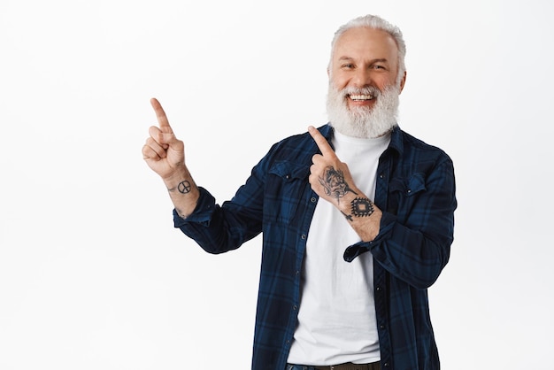 Hombre mayor feliz con tatuajes apuntando al logo de la esquina superior izquierda riendo alegre de pie contra fondo blanco Copiar espacio