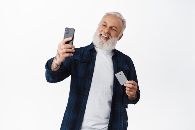 Hombre mayor feliz tomando selfie con su tarjeta de crédito, sonriendo mientras paga en línea con identificación facial en la aplicación de teléfono inteligente, comprando en la tienda de Internet, de pie contra la pared blanca