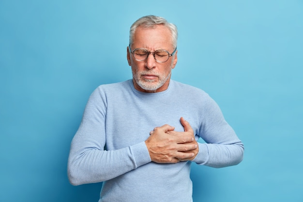 Hombre mayor con gafas presiona la mano contra el pecho tiene un ataque al corazón sufre de dolor insoportable cierra los ojos usa gafas ópticas posa contra la pared azul