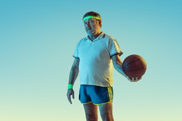 Hombre mayor jugando baloncesto sobre fondo degradado en luz de neón. Modelo masculino caucásico en gran forma se mantiene activo, deportivo.