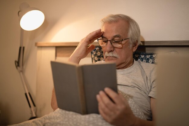 Hombre mayor leyendo un libro mientras se acuesta en la cama por la noche