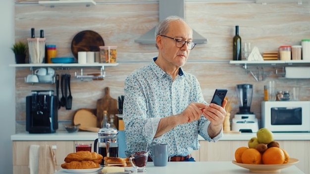 Hombre mayor navegando en internet usando un teléfono inteligente en la cocina mientras disfruta del café de la mañana durante el desayuno. Auténtico retrato de jubilados disfrutando de la moderna tecnología en línea de Internet