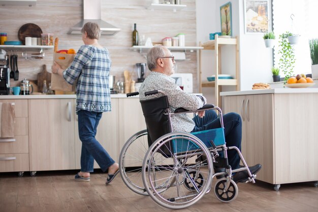 El hombre mayor no válido que sonríe mirando por la ventana en la cocina y la esposa está desempacando comestibles. Inválido, pensionista, minusválido, parálisis.