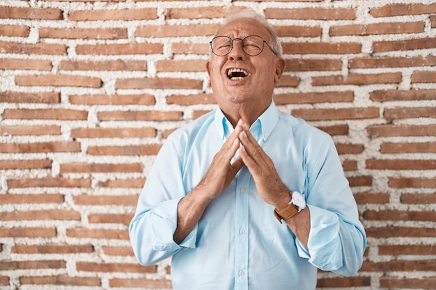 Foto gratuita hombre mayor con pelo gris parado sobre una pared de ladrillos mendigando y orando con las manos junto con expresión de esperanza en la cara muy emocional y preocupada mendicidad