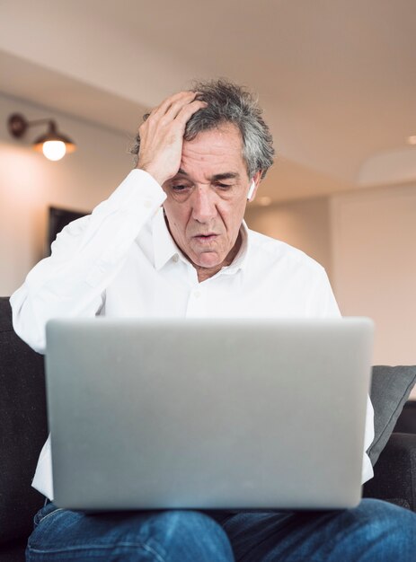 Hombre mayor preocupado que se sienta en el sofá que mira la computadora portátil