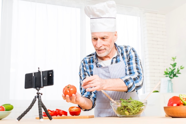 Hombre mayor que hace videollamada en el teléfono móvil que muestra el tomate de la herencia mientras que prepara la ensalada