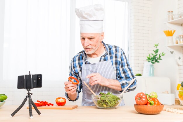 El hombre mayor que lleva el sombrero del cocinero blanco que hace videollamada mientras que cocina el alimento en la cocina