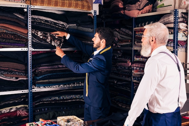 Foto gratuita hombre mayor que mira al sastre de sexo masculino que toma la tela del estante en el taller