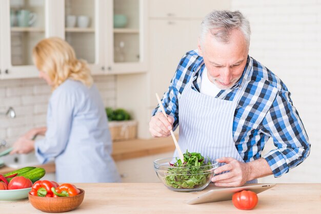 Hombre mayor que mira la tableta digital que prepara la ensalada verde en el bol de vidrio y su esposa que trabaja en el fondo