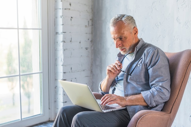 Hombre mayor que se sienta en el sillón que usa la computadora portátil