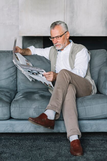 Hombre mayor que se sienta en el sofá acogedor con el periódico de la lectura de la pierna cruzada