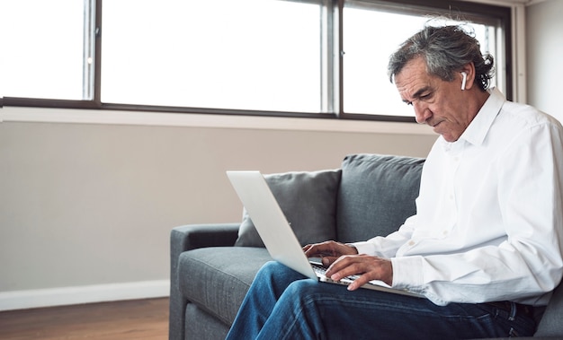 Hombre mayor que se sienta en el sofá usando la computadora portátil